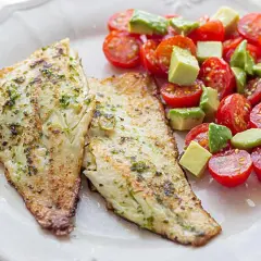Dos filetes de lubina a la plancha, dorados y con hierbas, acompañados de una ensalada fresca de tomates cherry y aguacate en cubos.