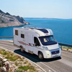 Un camping-car Adria roule sur une route côtière en Sardaigne, avec la mer bleue et des montagnes rocheuses en arrière-plan.