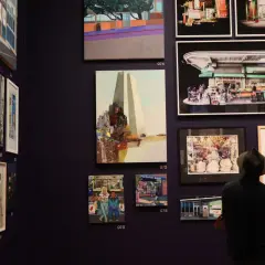 A man in a hat views diverse artworks in a gallery, showcasing vibrant cityscapes and street scenes, typical of young art galleries.