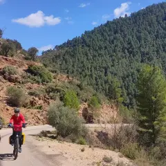 Dos ciclistas exploran rutas gravel por un camino rural rodeado de montañas boscosas y terreno rocoso bajo un cielo azul.