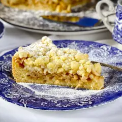 Ein Stück von Omas Apfelkuchen mit Streuseln auf einem blauen Teller, bestäubt mit Puderzucker.