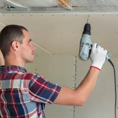 Mężczyzna w rękawiczkach montuje uchwyt do sufitu podwieszanego za pomocą wkrętarki.