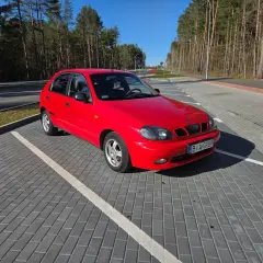 Czerwone Daewoo Lanos na parkingu z kostki brukowej, w tle las i błękitne niebo.