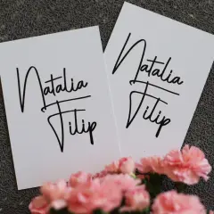 Dwie białe kartki z napisem "Natalia Filip" w ozdobnej czcionce, na tle szarej tekstury i różowych kwiatów.