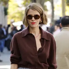 Mujer con corte francés con raya lateral, gafas oscuras y labial rojo, luciendo un suéter marrón.