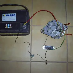 Akumulator Varta, alternator, regulator napięcia i żarówka połączone przewodami na podłodze.