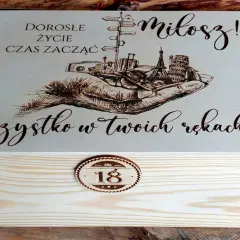 Drewniana skrzynka z grawerem na 18 urodziny od chrzestnej. Na wieczku napis "Dorosłe życie czas zacząć, Miłosz! Wszystko w Twoich rękach!" i grafika dłoni z symbolami podróży.