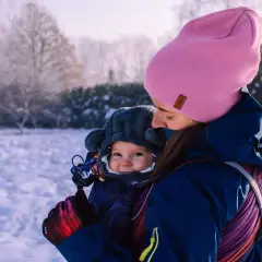 Mama w różowej czapce i granatowej kurtce tuli dziecko w nosidełku. Zima w pełni, śnieg wokół. Jak ubrać dziecko do nosidełka zimą? Warstwowo i ciepło!