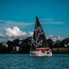Żaglówka z czarnym żaglem z białym wzorem płynie po jeziorze. Załoga na pokładzie, w tle drzewa i domy. To piękna regata.