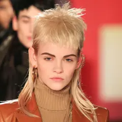 Ein Model präsentiert einen modernen Mullet mit blonden Locken, auffälligen Ohrringen und einem braunen Rollkragenpullover.