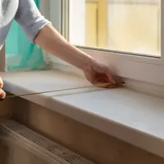 Mierzenie parapetu zlicowanego ze ścianą. Dłoń trzyma taśmę mierniczą, druga dłoń przytrzymuje koniec przy oknie.