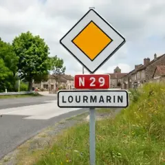 Panneau "je suis prioritaire" à l'entrée de LOURMARIN sur la N29. Le village s'étend derrière, bordé d'arbres et de maisons anciennes.
