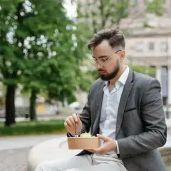 Mężczyzna w okularach i marynarce robi sobie przerwę w pracy, jedząc sałatkę na ławce w parku.