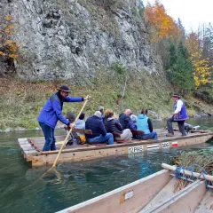 Flisacy na tratwie płyną Dunajcem, jesienne drzewa i skalne zbocza tworzą malowniczą scenerię. Przystań flisacka wita turystów.