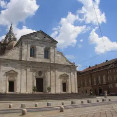 Il Duomo di Torino, con la sua imponente facciata in pietra e la cupola maestosa, si staglia sotto un cielo azzurro punteggiato di nuvole bianche.
