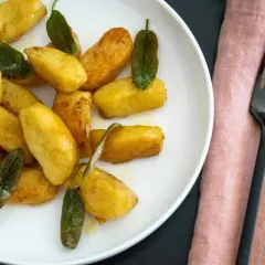Goldbraune salbei gnocchi auf weißem Teller, garniert mit frischen Salbeiblättern. Daneben liegen schwarzes Besteck auf rosafarbenem Stoff.