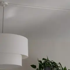 Lámpara de techo con un punto de luz desplazado, colgada de un riel blanco. Una planta verde asoma por la derecha.