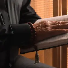 Dłoń starszego mężczyzny spoczywa na otwartej Biblii. Wokół nadgarstka ma drewniane paciorki. Gdzie jest pastor?