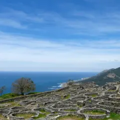 Ruinas circulares de piedra en el mirador Santa Tecla, Galicia, con el mar azul y montañas verdes al fondo.