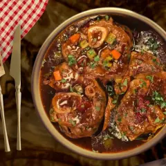 Un plat d'osso bucco de veau traditionnel, mijoté dans une sauce riche, garni de persil frais et de légumes. Servi avec une serviette à carreaux rouges et blancs et des couverts.