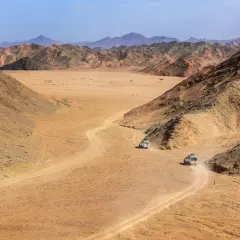 Pustynny krajobraz z krętą drogą, po której jadą dwa samochody terenowe. Idealne miejsce na **marsa alam wycieczki fakultatywne**.