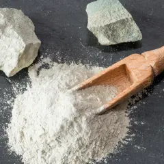 Feiner Pulver aus Magnesium-Aluminium-Silikat wird mit einer Holzschaufel auf dunklem Stein verteilt, daneben liegen zwei Brocken des Materials.