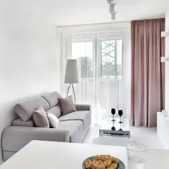 Jasny, bardzo mały salon w bloku z szarą kanapą, stolikiem kawowym i widokiem na balkon. Obok widać łóżko.