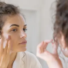 Woman applying cream to her face in a mirror.