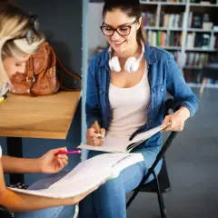 Dwie studentki uczą się do egzaminów w bibliotece. Jedna z nich, w okularach i dżinsowej koszuli, uśmiecha się, pisząc w zeszycie. To idealne miejsce do nauki w **liceum dla dorosłych 2-letnim**.