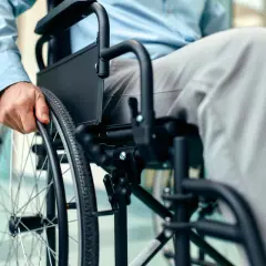 Osoba w jasnej koszuli i szarych spodniach siedzi na wózku inwalidzkim, trzymając ręką obręcz koła.