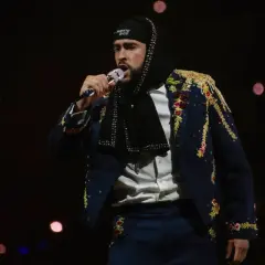 Bad Bunny w ozdobnej marynarce śpiewa na koncercie na ciemnym tle z rozmazanymi światłami.