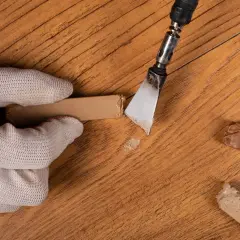 Guanto bianco rimuove residui con spatola, mostrando come togliere graffi dal parquet. Riparazione fai-da-te.