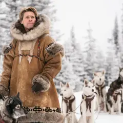 Man i pälsrock med hundspann i snöig skog. Scen från en togo film.