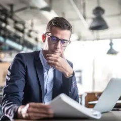 Mężczyzna w okularach i garniturze analizuje dokumenty przy laptopie w kawiarni.