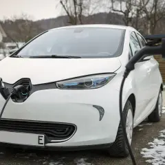 Białe auta elektryczne podłączone do ładowarki na tle domu i drzew.