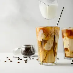 Versare latte freddo in un bicchiere di caffè ghiacciato crea un effetto marmorizzato invitante. Chicchi di caffè sparsi completano la scena.