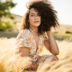 A woman with curly hair poses in a field of tall grass, her dress catching the golden hour light. This image demonstrates how to shoot with a flash for a soft, natural look.