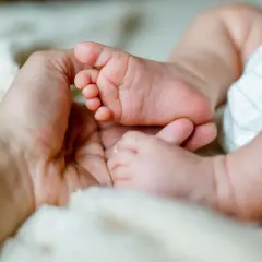 Dłoń rodzica trzyma stópkę niemowlęcia na miękkim kocyku.