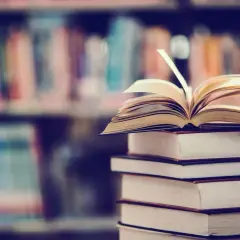Stos książek, na wierzchu otwarta księga. W tle regały pełne literatury, w tym podręczniki do prawa pracy.