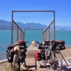 Dos bicicletas equipadas para aventura esperan junto a un muelle en la Carretera Austral. El agua turquesa y las montañas azules crean un paisaje impresionante.