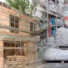 Budowa domu w toku, obok magazyn materiałów budowlanych. Widać palety z cementem i półki z różnymi produktami. Ceny materiałów budowlanych wpływają na koszty inwestycji.