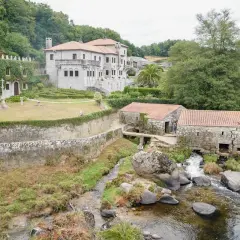 Antigua barcala con molinos de piedra junto a un río caudaloso, con una casa señorial cubierta de hiedra al fondo.