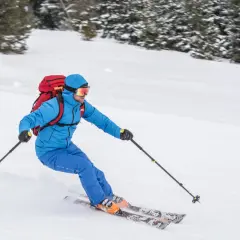 Narciarz w niebieskiej kurtce i goglach zjeżdża po ośnieżonym stoku, niosąc czerwony plecak. To prawdziwy miłośnik ski tury!