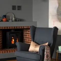 Mysigt vardagsrum med grå väggfärg, en bekväm fåtölj framför en eldstad och en tavla.