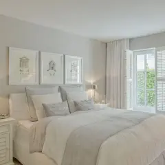 Helles, luftiges Schlafzimmer im Strandhaus-Stil mit weißem Bett, Kissen und Gardinen. Drei gerahmte Zeichnungen über dem Bett.
