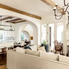 A cozy living room with a fireplace and kitchen, featuring exposed wooden beams and arched doorways, embodying a warm mediterranean interior design.
