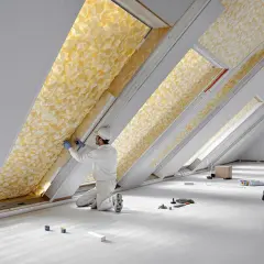 Un ouvrier installe des matériaux isolants jaunes texturés dans un grenier. L'isolation est visible dans plusieurs lucarnes.