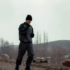 A boxer in a black tracksuit and beanie is running outdoors, practicing his footwork. The barren landscape suggests a cold day for this intense training.