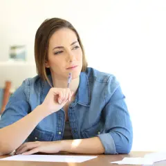 Femme songeant à une lettre de remise gracieuse impot taxe d'habitation, stylo à la main, devant des papiers.