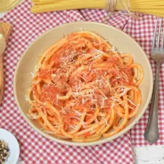 Miseczka pełna aromatycznego makaronu amatriciana, posypanego startym parmezanem. Obok kawałek sera, ziarna pieprzu i widelec.
